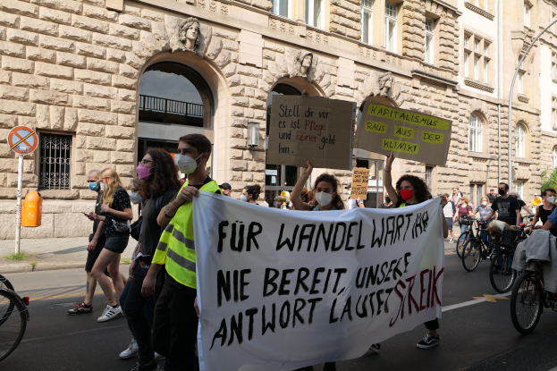 Eine Gruppe von Menschen marschiert auf einer Straße in Berlin, hält Schilder und Banner hoch und fährt dabei Fahrräder, vor einem Gebäude mit Bögen, Säulen, Skulpturen und Bäumen.