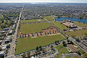 Luftaufnahme einer Stadt mit Gebäuden, Bäumen, Gras, Straßen mit Fahrzeugen, Wasser und bewölktem Himmel, die ein zentrales Feld zeigt, das als Parkplatz vorgeschlagen ist.