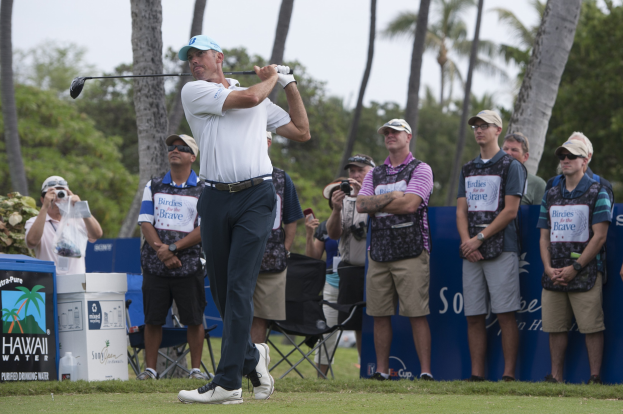 Ein Mann in einem weißen T-Shirt, schwarzen Hosen und einer blauen Mütze schwingt einen Golfschläger auf einem grünen Feld, mit Zuschauern und Fotografen dahinter, vor Bäumen und einem klaren blauen Himmel.
