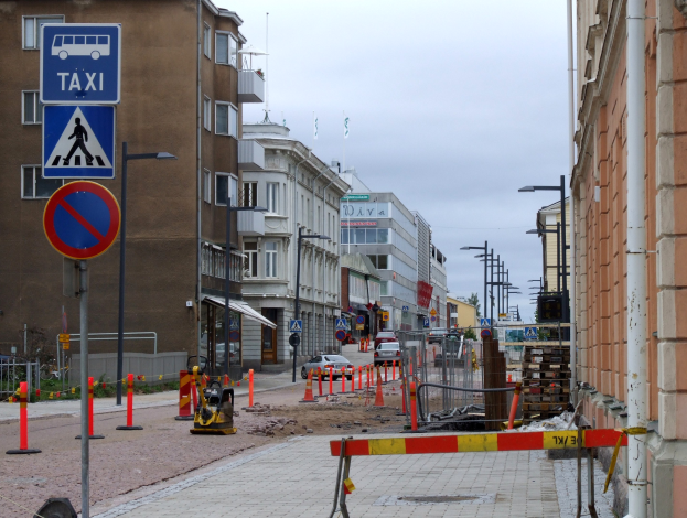 Eine Stadtstraße mit Gebäuden, Straßenlaternen, Verkehrszeichen, Verkehrskegeln, Kraftfahrzeugen, Absperrpoller, Bäumen, einer Baustelle mit Verkehrsschildern und einem bewölkten Himmel im Hintergrund.