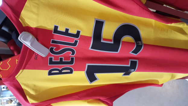 Ein rotes und gelbes Fußballtrikot mit aufgedrucktem Text und Nummern, das an einer Ladengestell hängend zu sehen ist, mit anderen Gegenständen und dem Boden im Hintergrund.