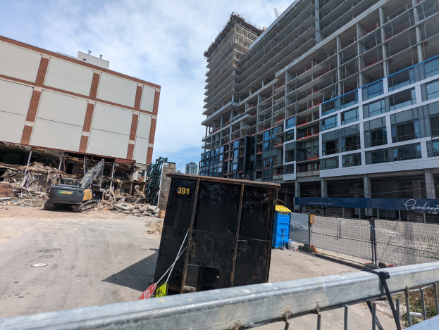 Großes Gebäude mit Schutt davor, umgeben von einem Metallzaun, Container, Pflanzen, Bäumen, Pfählen, einem Schild, Fahrzeugen, Steinen und einem bewölkten Himmel, was auf eine Baustelle für einen neuen Wohnkomplex hinweist.