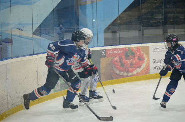 Gruppe junger Menschen, die Eis hockey auf einer Indoor-Eisbahn spielen, mit Helmen, Sportuniformen und Schlittschuhen, während sie Hockey-Schläger halten, mit einem Plakat im Hintergrund auf der Glaswand.