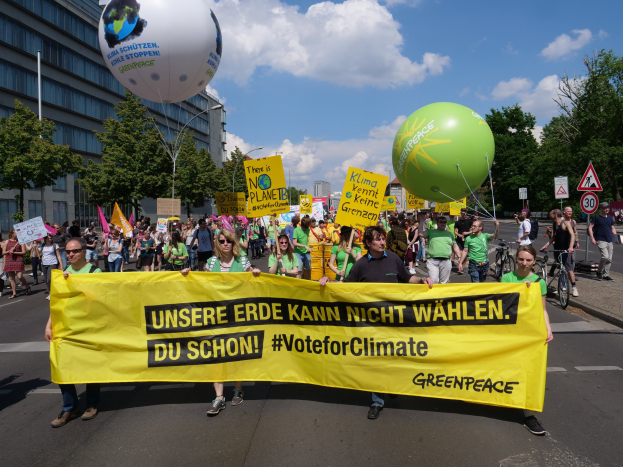 Eine Gruppe von Menschen marschiert auf einer Straße und hält ein gelbes 'Wähle für den Klimaschutz'-Plakat hoch, mit Ballons, Schildern, Fahrrädern, Bäumen, Laternenpfählen und Gebäuden im Hintergrund und einem klaren blauen Himmel.