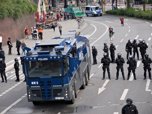 Gruppe von Polizeibeamten vor einem gepanzerten Fahrzeug auf einer Stra├če mit bewaffneten Individuen und urbaner Umgebung, einschließlich Geb├Ąuden, Verkehrszeichen und Vegetation.