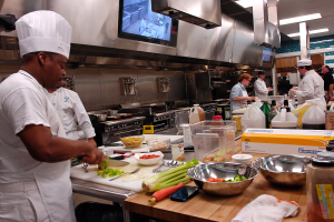 Eine Gruppe von Köchen in einer Küche, die Essen auf einem Schneidbrett zubereiten, wobei ein Koch im Vordergrund in einem weißen Kleid und einer Mütze steht und ein Messer hält und von Utensilien, Schüsseln, Gemüse, einem Mobiltelefon, Flaschen und anderen Gegenständen umgeben ist, während im Hintergrund ein Fernseher und Deckenleuchten zu sehen sind.