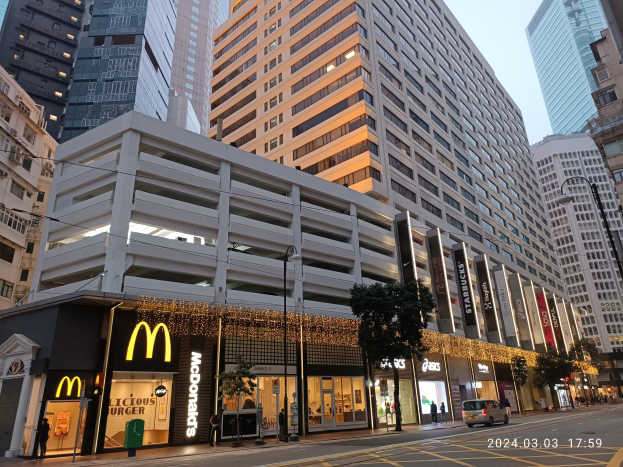 Ein McDonald's-Restaurant auf einer belebten Stadtstraße mit hohen Gebäuden, Fahrzeugen, Fußgängern, Straßenmöbeln, Bäumen und einem klaren Himmel.