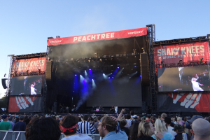Eine große Menschenmenge steht vor einer hell erleuchteten Bühne beim Shaky Knees Music Festival, auf der Musiker unter Textbannern spielen, mit einem Geländer im Vordergrund und einem klaren blauen Himmel im Hintergrund.