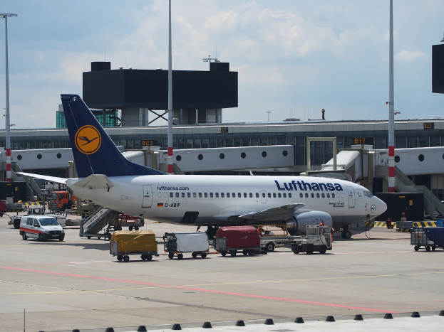 Lufthansa Airbus A320-200 am Frankfurter Flughafen mit Bodenfahrzeugen und Wagen vorne, einem Gebäude und Laternen im Hintergrund unter einem klaren blauen Himmel.
