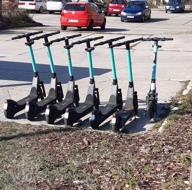 Eine Reihe von Elektro-Scootern, die auf der Seite einer Straße geparkt sind, mit Gras und trockenen Blättern, Autos, Bäumen, Gebäuden und einem klaren blauen Himmel im Hintergrund.