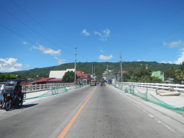 Autobahn mit fahrenden Fahrzeugen, ein geparktes Motorrad auf dem Seitenstreifen, Geländer, Strommäste mit Drähten, Bäume, Gebäude, entfernte Hügel und ein bewölkter Himmel.