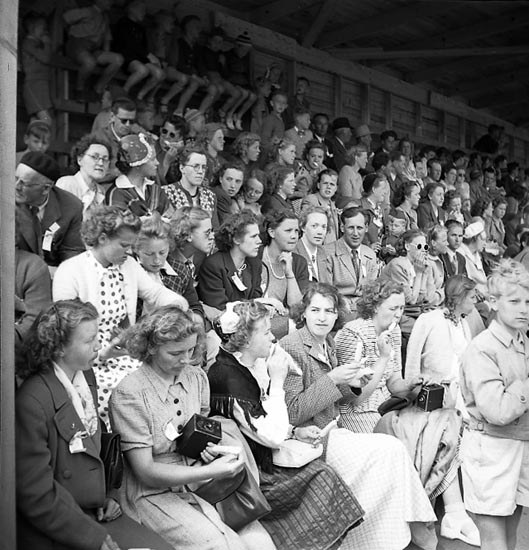 Schwarzes und weißes Foto einer Menge, die in einem Stadion sitzt und ein Sportereignis beobachtet, mit einer hölzernen Wand und einem Dach im Hintergrund.