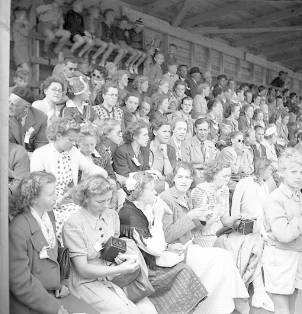 Schwarzes und weißes Foto einer Menge, die in einem Stadion sitzt und ein Sportereignis beobachtet, mit einer hölzernen Wand und einem Dach im Hintergrund.