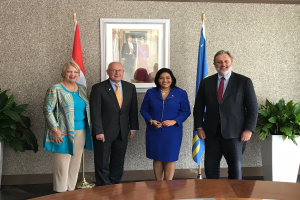 Eine Gruppe strahlender Menschen um einen Tisch mit Papieren herumstehend, umgeben von Fahnen, Topfpflanzen und einem Fotorahmen an der Wand, feiert die Unterzeichnung eines Memorandums zwischen den Vereinigten Staaten und Kanada.