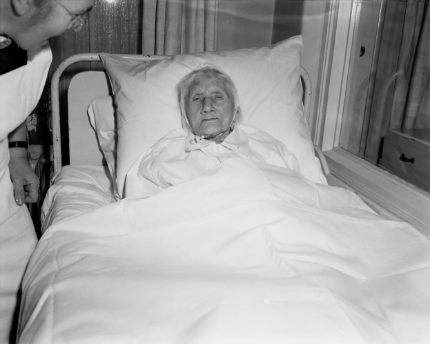 Ein älteres Weib liegt in einem Krankenhausbett mit einer weißen Decke und einem Kissen, während eine Krankenschwester neben ihr steht, mit einem Fenster mit Vorhängen im Hintergrund.