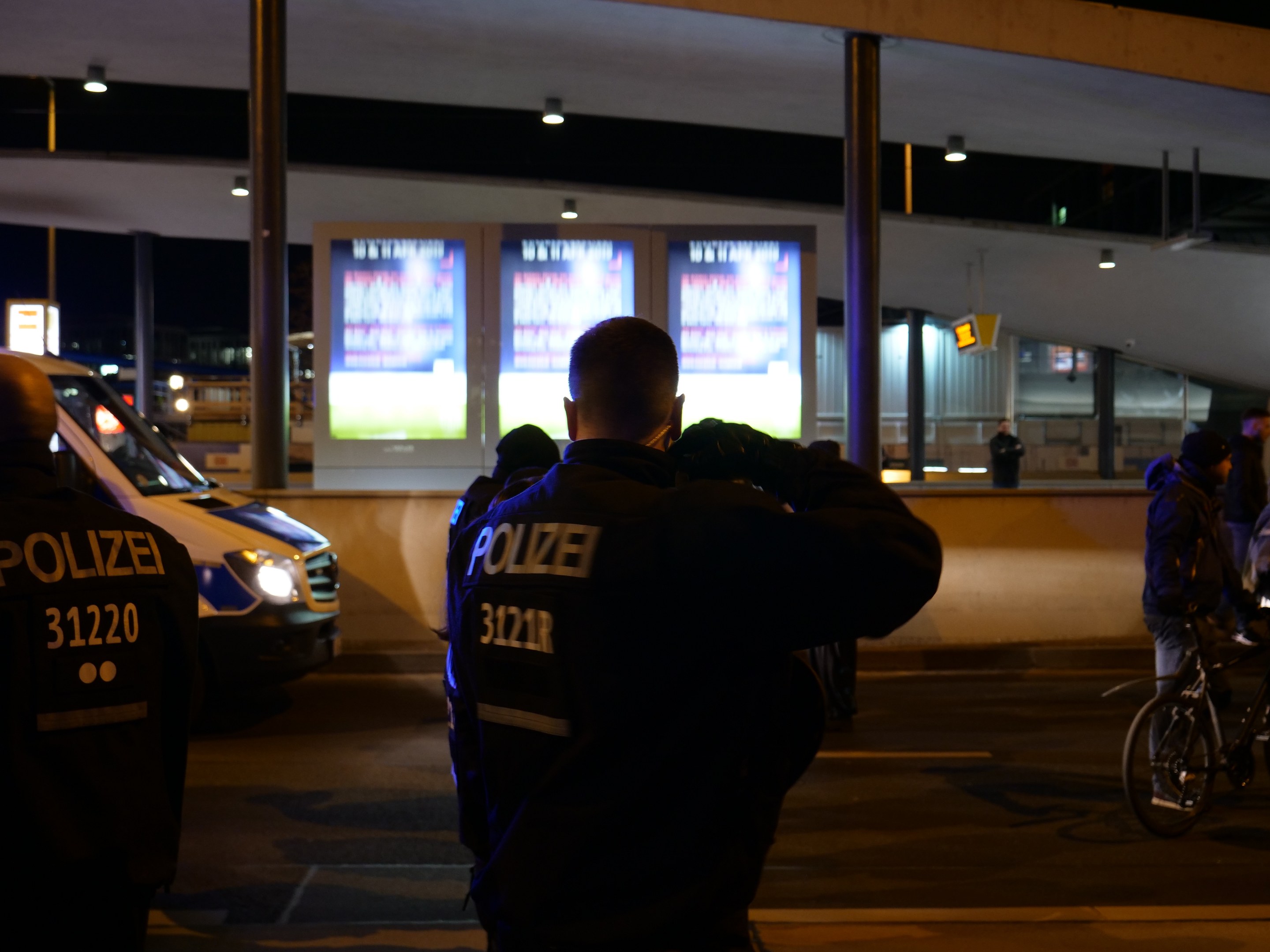 Eine Gruppe von Polizisten steht vor einem Gebäude in der Nacht mit einem geparkten Auto auf der linken Seite und einem Radfahrer auf der rechten Seite, beleuchtet von Bildschirmen und Lichtern im Hintergrund.