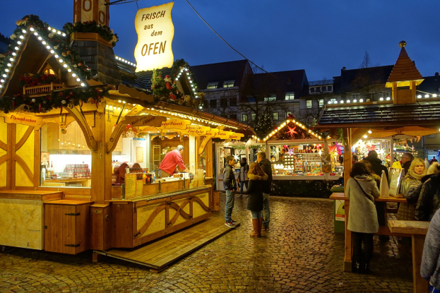 Ein lebendiger Weihnachtsmarkt auf einem Kopfsteinpflasterweg bei Nacht, mit Menschen, die an beleuchteten Ständen und festlichen Dekorationen vorbeischlendern, vor Buildings, Bäumen und einem klaren Himmel.