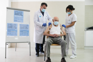 Ein älterer Mann sitzt in einem Krankenhauszimmer auf einem Stuhl, während ihm zwei maskierte und handschuhgetragene Ärzte eine Impfung verabreichen, ein Schild mit Text auf der linken Seite und Gegenstände auf einer Plattform auf der rechten Seite.