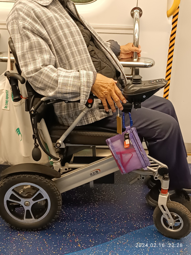 Ein älterer Mann im Rollstuhl in einem Zug, mit einer Tasche neben ihm und einem Spiegel an der Wand hinter ihm.