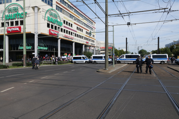 Polizisten stehen auf der Straße neben einer Tram, mit Fahrzeugen, Strommasten, Gebäuden, Bäumen und einem bewölkten Himmel im Hintergrund.