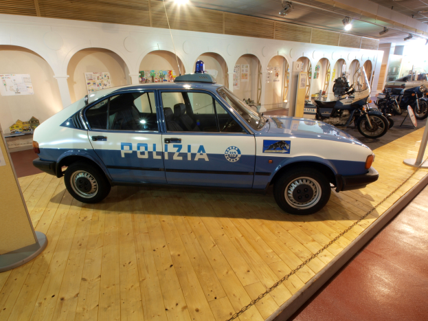 Ein Polizeiwagen wird in einem Museum ausgestellt, umgeben von Motorrädern und anderen Fahrzeugen, mit einer Säule und einer Tafel auf der linken Seite, Plakaten an der Hintergrundwand und Dachleuchten, die die Szene beleuchten.