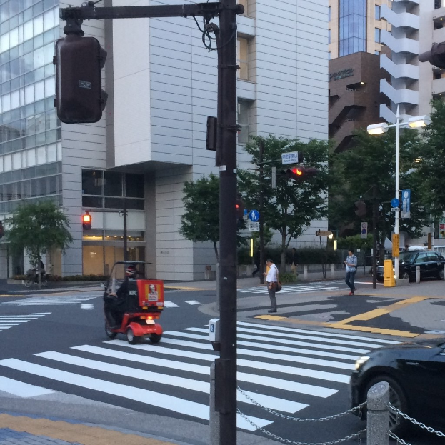 Ein E-Scooter auf einer Straße mit Personen im Hintergrund, eine Ampel im Vordergrund und Gebäude mit Bäumen im Hintergrund.