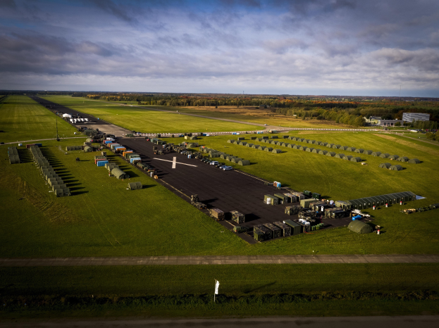 Luftaufnahme einer Militärbasis in den Niederlanden mit Fahrzeugen, Rasenflächen, Bäumen, Gebäuden und einem bewölkten Himmel.