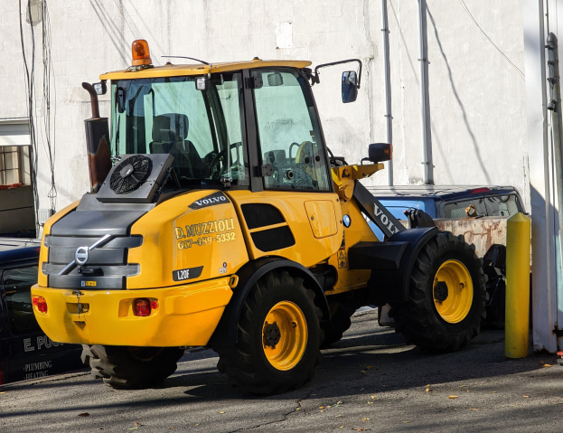 Ein gelber Radlader steht auf einer Straße vor einem Gebäude mit einer Wand im Hintergrund, umgeben von anderen Fahrzeugen.