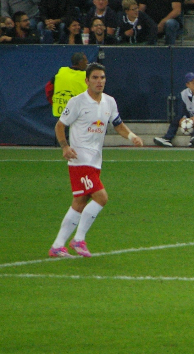 Ein Mann in weißem Hemd und roter Hose, der auf einem Feld Fußball spielt, umgeben von einer Gruppe von Menschen im Hintergrund.