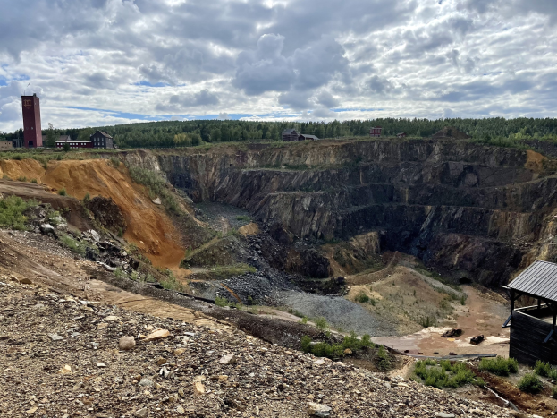 Großer Tagebau auf einem Schmutzfeld umgeben von Bäumen, Gebäuden und einem Schuppen unter einem bewölkten Himmel, mit verstreuten Steinen und Pflanzen.