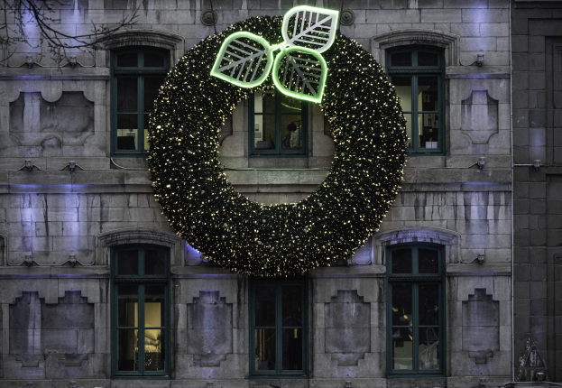 Ein festlicher Adventskranz aus beleuchtetem Efeu hängt an der Seite eines Gebäudes mit Fenstern und einem Baum links daneben.