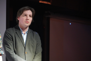 Zwei Personen stehen nebeneinander auf einer Bühne, eine in einem Anzug und die andere neben ihnen, beide mit ernsten Gesichtern.