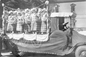 Schwarzes und weißes Bild einer Gruppe von Frauen, die auf einem Lkw stehen und Körbe halten, mit einem Gebäude und Bäumen im Hintergrund.