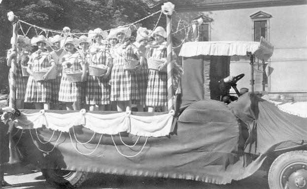 Schwarzes und weißes Bild einer Gruppe von Frauen, die auf einem Lkw stehen und Körbe halten, mit einem Gebäude und Bäumen im Hintergrund.