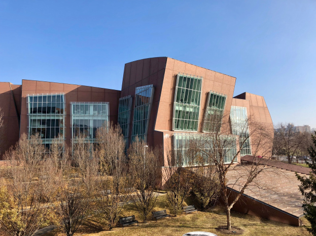 Großes Gebäude mit vielen Fenstern, umgeben von Bäumen, Bänken, Gras und einem Laternenmast, mit anderen Gebäuden und einem klaren blauen Himmel im Hintergrund; Teil des neuen Campus der University of Illinois at Chicago.