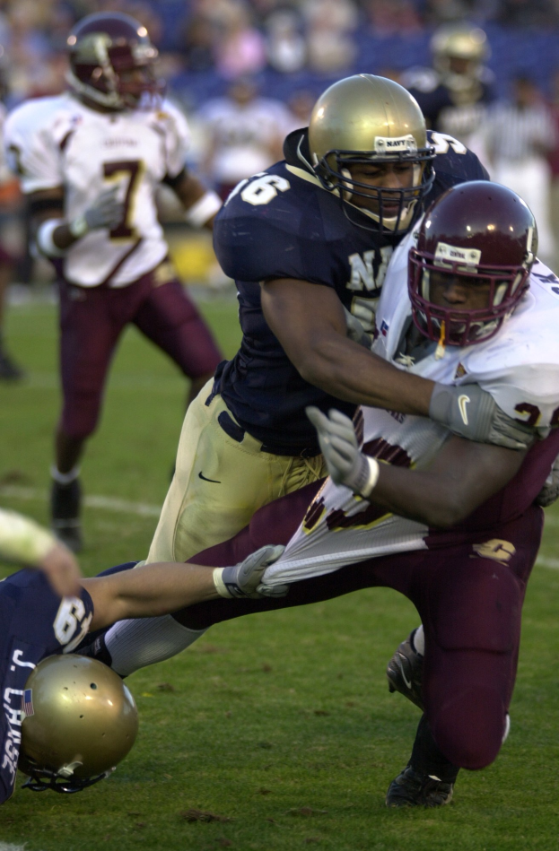 Zwei Fußballspieler in Helmen und Uniformen kämpfen auf dem Feld, während andere zusehen.