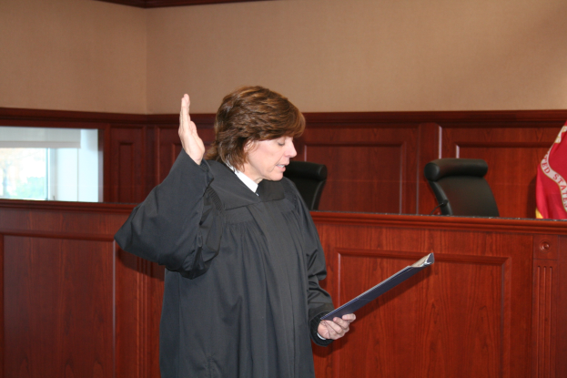 Eine Frau in schwarzer Robe steht vor einem Richtertisch, hält eine Akte, mit Mikrofon und Flagge auf dem Tisch, Stühle dahinter und eine fensterumrandete Wand im Hintergrund.
