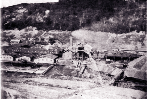 Schwarzes und wei├čes Foto eines Kohlebergbaus mit Geb├Ąuden, Masten und B├Ąumen im Hintergrund, mit Text unten.