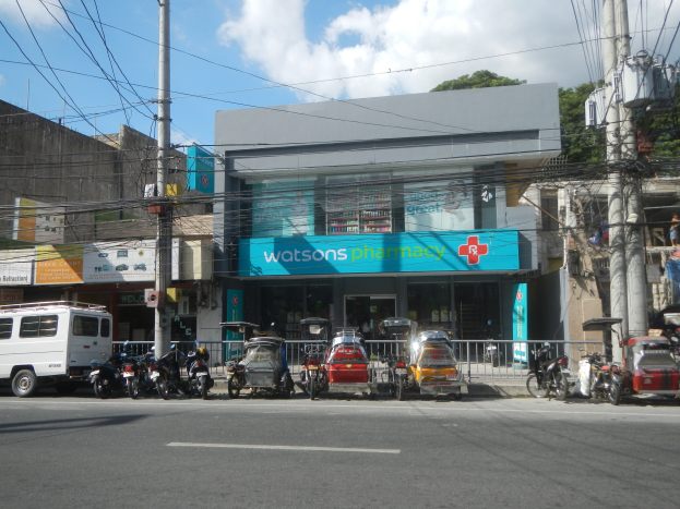 Eine belebte Stadtstraße mit Fahrzeugen, Strommasten mit Drähten, Gebäuden einschließlich einer Apotheke mit einem vor der Tür geparkten Lieferwagen, Bäumen und einem bewölkten Himmel.