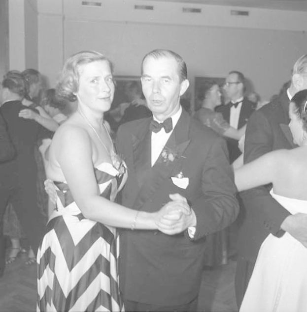 Zwei Menschen, ein Mann in einem schwarzen Blazer und weißem Hemd mit schwarzer Hose und eine Frau in einem unbekannten Outfit tanzen zusammen auf einer formellen Veranstaltung, halten sich an den Händen und wiegen sich im Takt gegen eine Wand im Hintergrund.