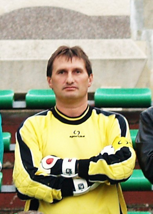 Ein Torwart in einem gelben Trikot steht neben einem Mann in einer schwarzen Jacke, mit grünen Stühlen und einer Wand im Hintergrund.