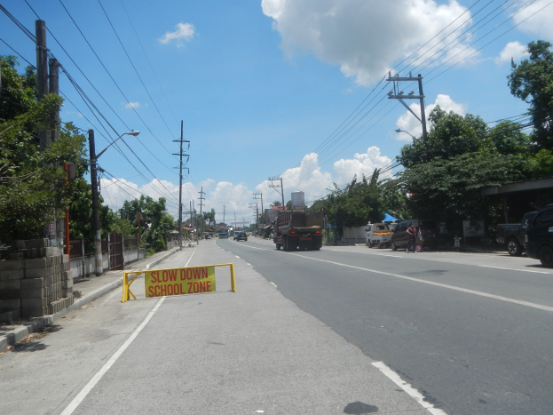 Eine Straße mit Fahrzeugen, ein "Slow Down School Zone"-Schild, Strommasten mit Drähten, Bäume, Straßenlaternen und ein bewölkter Himmel.