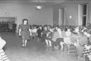 Eine Frau in einem schwarzen und weißen Kleid läuft über den Laufsteg während einer Modenschau für Frauenwahlrecht, mit einer Menge, die auf Stühlen sitzt und einer Uhr und Lampen an den umliegenden Wänden.