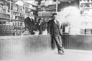 Drei Männer stehen an einem Tresen in einem Laden, umgeben von verschiedenen Gegenständen, mit einem Regal voller Bücher und anderen Gegenständen im Hintergrund, scheinbar an einem Raub beteiligt.