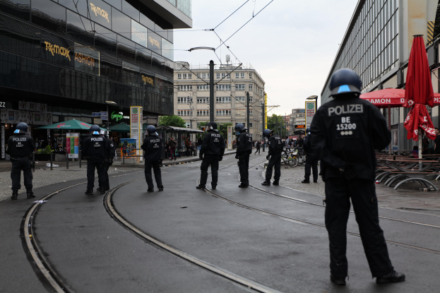 Eine Gruppe von Polizisten in schwarzen Uniformen und Helmen steht auf einer von Glasfenstern gesäumten Straße, mit Bäumen, Pfählen und verstreuter Außenmöblierung im Hintergrund unter einem klaren Himmel.