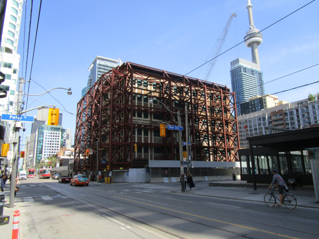 Eine belebte Stadtstraße mit einem großen Baugerüst im Hintergrund, mit Fahrzeugen, einem Radfahrer, Füßgängern, Ampeln, Masten mit Schildern, einem Kran und dem Himmel.