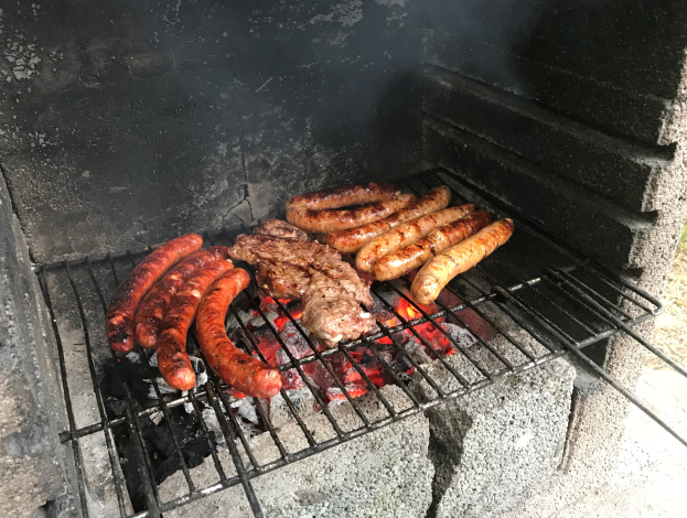 Grill mit Würstchen und Fleisch, das über Kohle und Feuer gegrillt wird, draußen auf Gras.