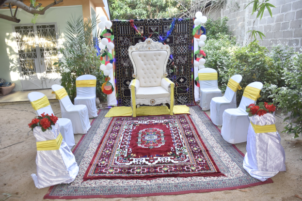 Eine Gartenhochzeitsaufstellung mit weißen Stühlen, einem roten Teppichläufer, Blumenbouquets, Dekorationsgegenständen, Luftballons, Topfpflanzen, Bäumen, einem Gebäude mit Fenstern und einer Tür und einer Wand auf der rechten Seite.