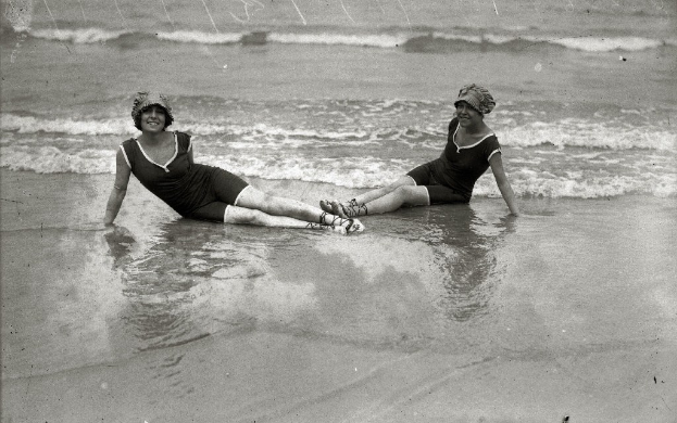 Zwei Frauen in Bikinis und Hüten sitzen im Meer, lächeln und genießen die Sonne und die Wellen auf einem Schwarz-Weiß-Foto.