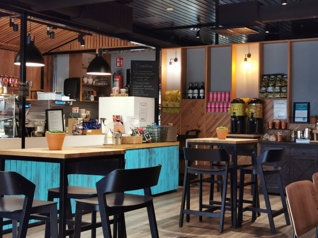 Innenansicht eines Restaurants mit Holztischen, Stühlen, einer Topfpflanze auf einem Tisch, einer Tafel, einem Kühlschrank, einem Feuerlöscher, Regalen mit Flaschen, Schränken mit Gegenständen, einem Foto auf der Wand, Deckenleuchten und Lampen.